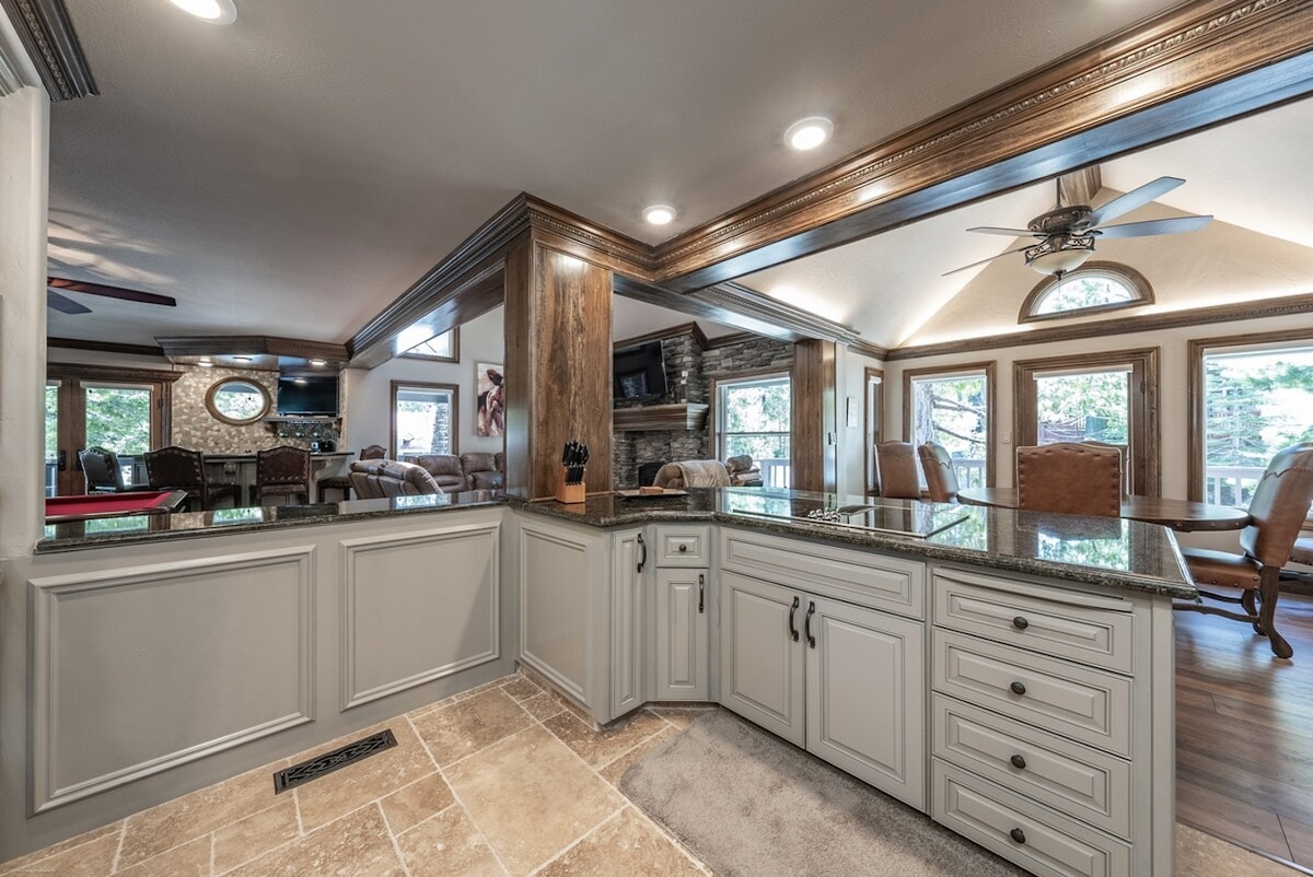 A spacious kitchen area is showcased with a center island featuring a dark granite countertop. Stylish cabinetry in light gray complements the open layout, which connects to a comfortable seating area visible in the background, illuminated by natural light from surrounding windows.