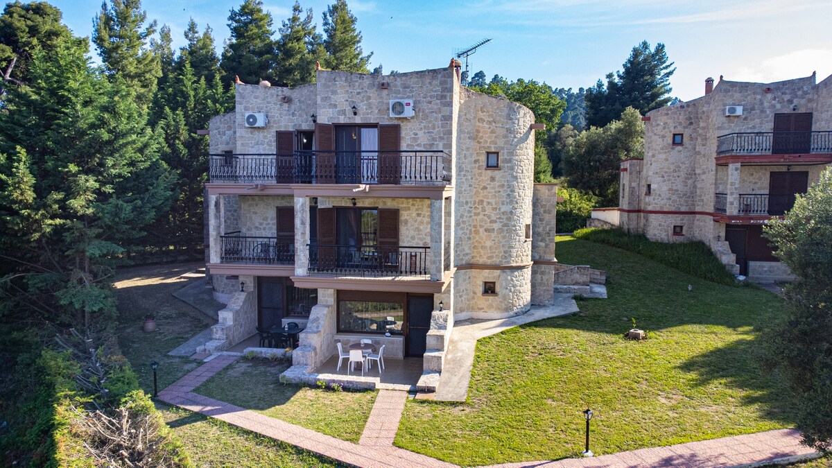 The exterior of a two-story stone house is presented, featuring balconies and air conditioning units. A grassy area surrounds the property, with pathways leading to the entrance. A small terrace with seating can be seen on the lower level, under a clear sky.