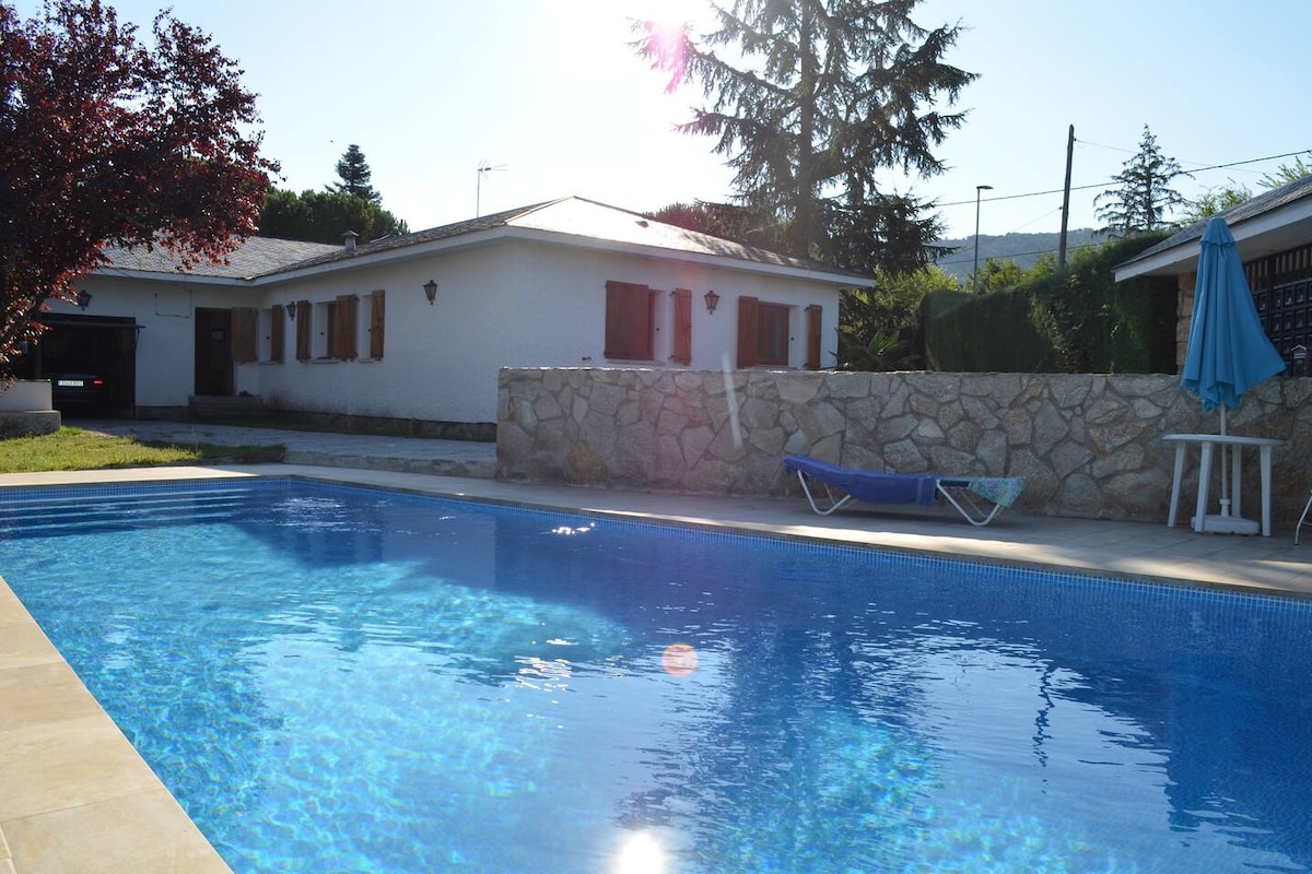 A large saltwater swimming pool reflects sunlight, surrounded by a paved terrace. A sun lounger and a blue umbrella provide relaxation space nearby. The rustic chalet is visible in the background, featuring wooden shutters and a stone wall that frames the pool area.