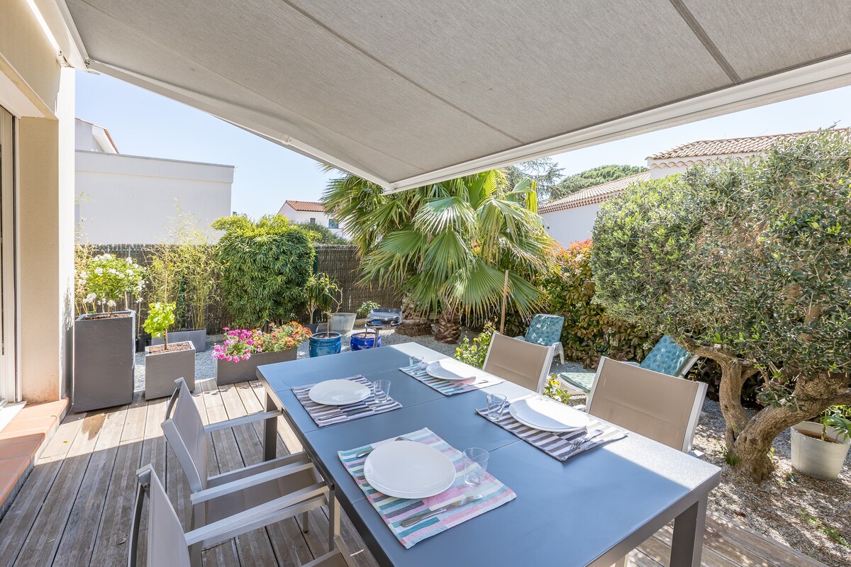 A shaded terrace is presented, featuring a dining table set with plates and stripes of fabric. Surrounding greenery, including palm trees and flowering plants, adds to the natural ambiance. A wooden deck complements the modern aesthetic of the outdoor space.
