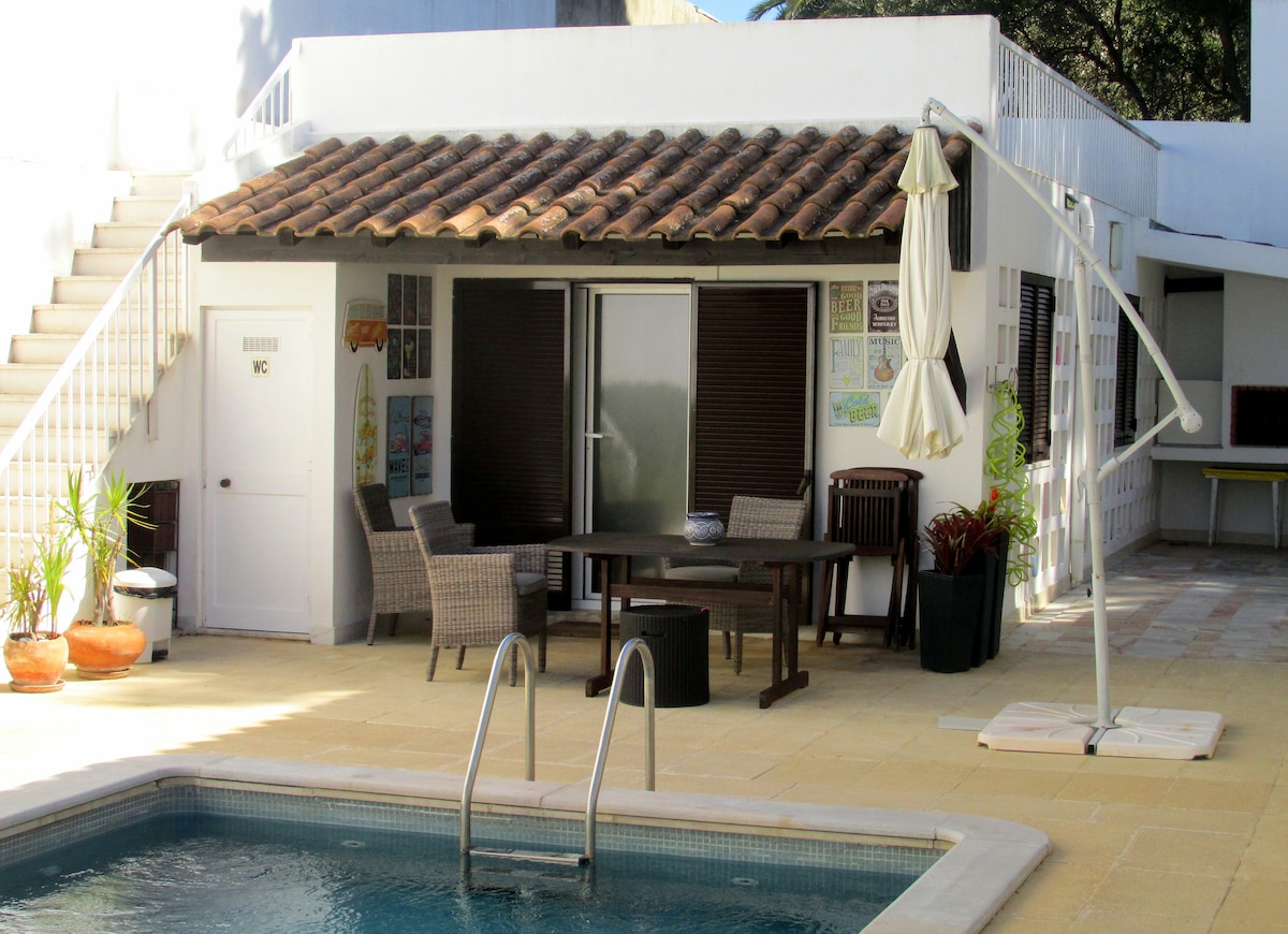 An outdoor area features a private swimming pool alongside a covered seating space. The seating area includes a table and chairs, with a large umbrella providing shade. Stairs lead to an upper level, and potted plants integrate greenery into the design.