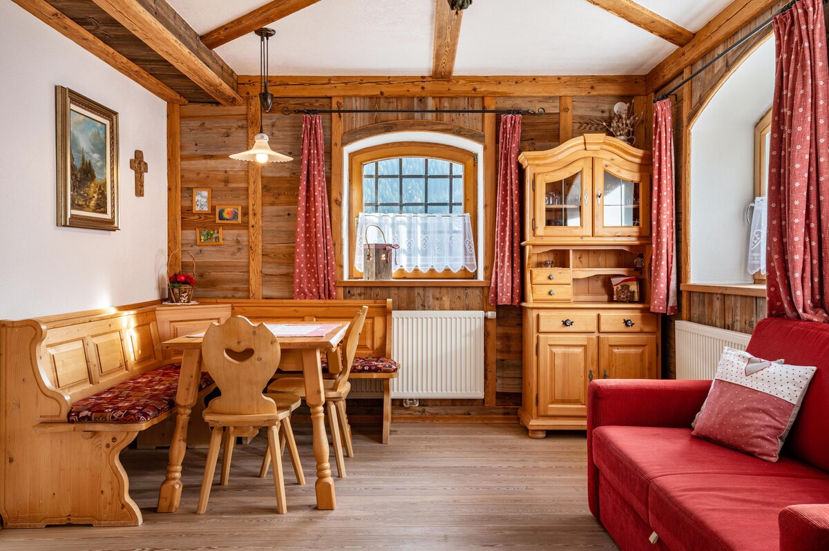 A wooden dining area is provided, featuring a corner bench and table, complemented by a collection of chairs. Natural light enters through a window with a decorative curtain. A sofa in warm red is positioned nearby, alongside a wooden sideboard adorned with decorative items.