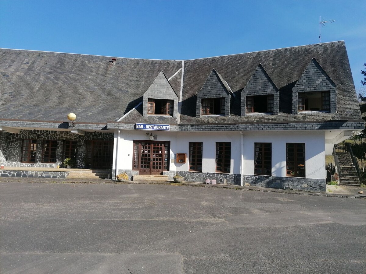 The hotel exterior is showcased, featuring a sloped roof with distinct dark shingles. The main entrance is framed by multiple windows, providing natural light. A sign indicating the restaurant is visible above the doors, with a stone facade complementing the white walls. Open outdoor space is prominent.