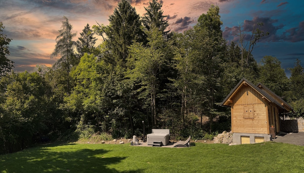 A spacious lawn area is bordered by lush trees, providing a private and serene ambiance. A modern hot tub sits on a stone terrace, inviting relaxation amidst nature. The wooden exterior of a nearby building complements the natural setting under a softly lit sky.
