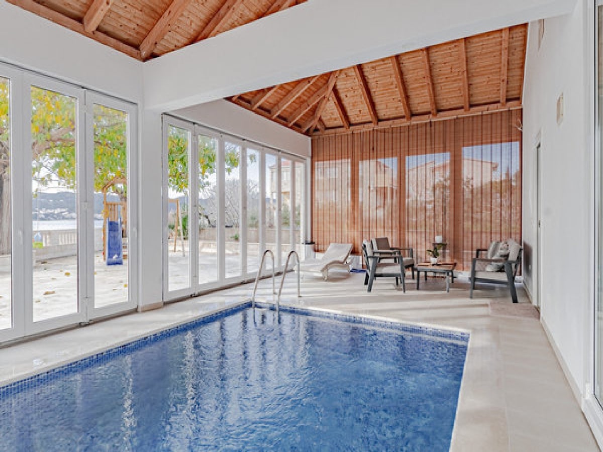 An indoor heated swimming pool is surrounded by large glass doors, allowing natural light to fill the space. Comfortable seating is available nearby, and wood-beamed ceilings add warmth to the area. Views of the outdoor landscape can be seen through the windows.