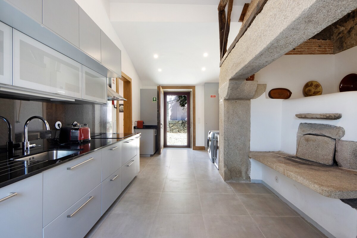 A modern kitchen features sleek cabinetry and a dark countertop. An ancestral fireplace is integrated into the stone wall near a cozy seating area. The space is illuminated by recessed lighting, offering a bright and functional environment.
