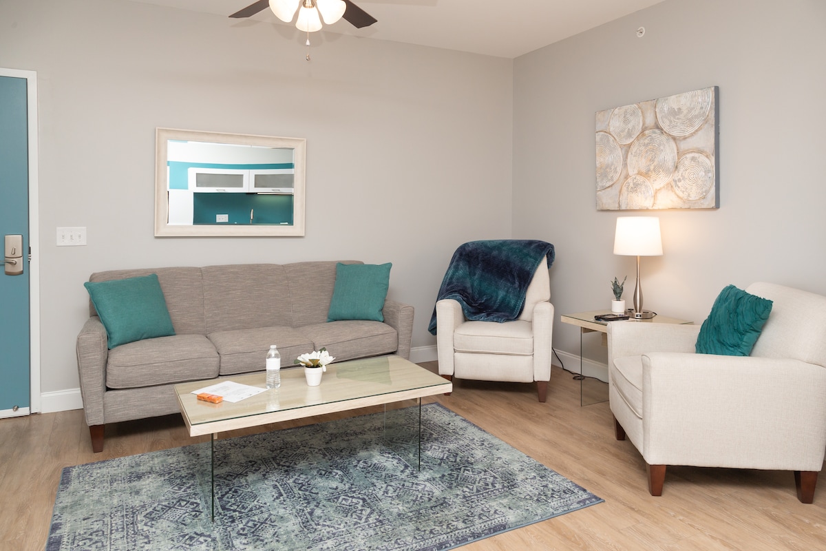 A comfortably arranged living area features a sofa adorned with teal cushions and two armchairs. A glass coffee table sits on a patterned rug, complemented by a small plant and a bottle of water. Light from a lamp creates a warm glow in the space.