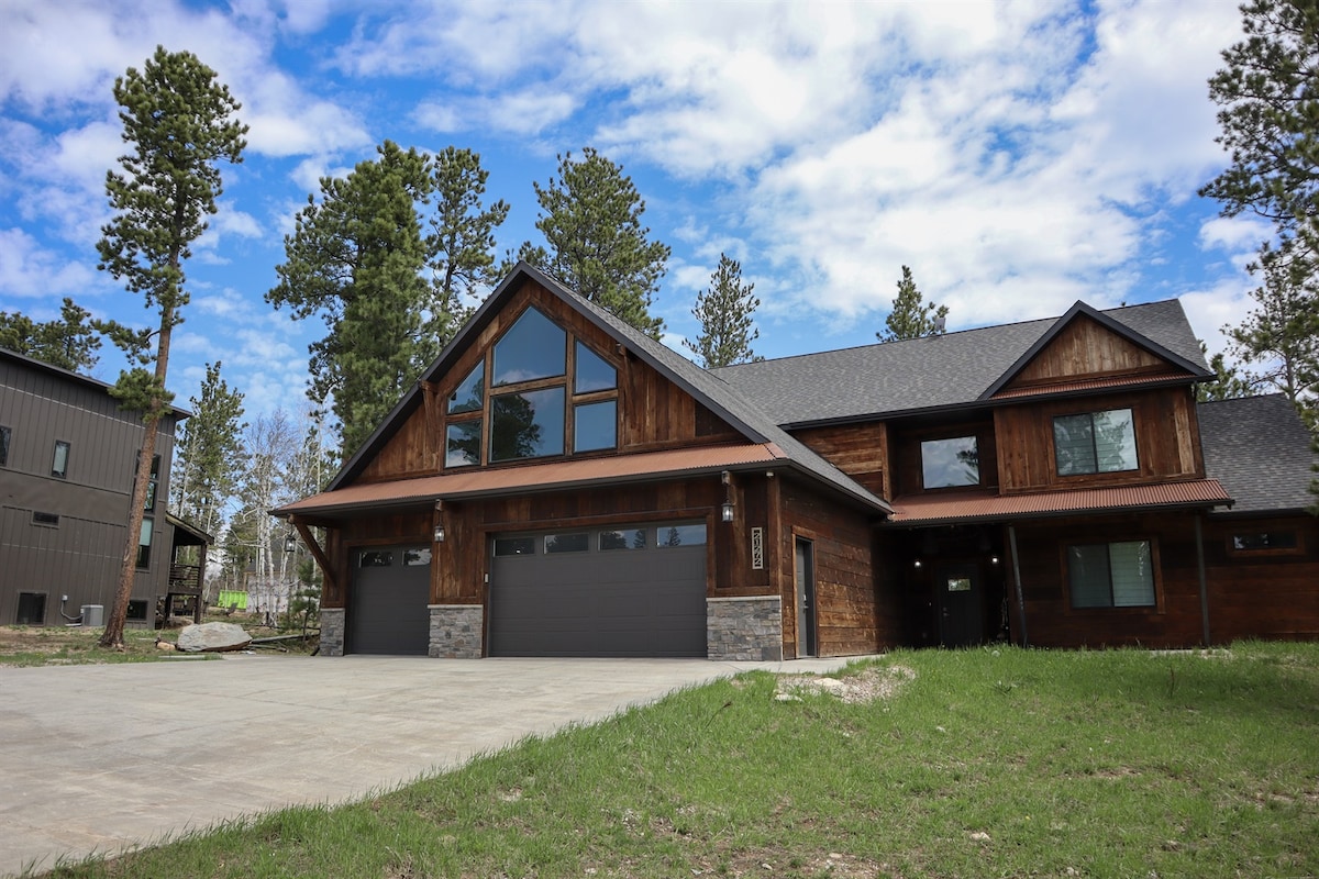 A modern, two-story cabin features a wooden exterior with large windows allowing natural light to fill the interior. The spacious driveway provides ample parking space, while surrounding greenery and tall pine trees create a serene backdrop.