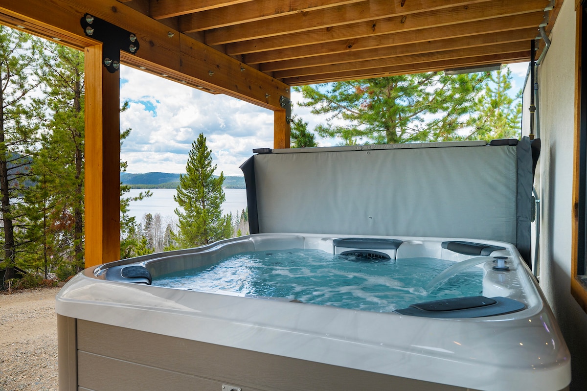 An outdoor hot tub is positioned under a wooden overhang, showcasing a serene view of the lake and surrounding pine trees. The bubbling water reflects the light from the sky, creating a calming atmosphere for relaxation.