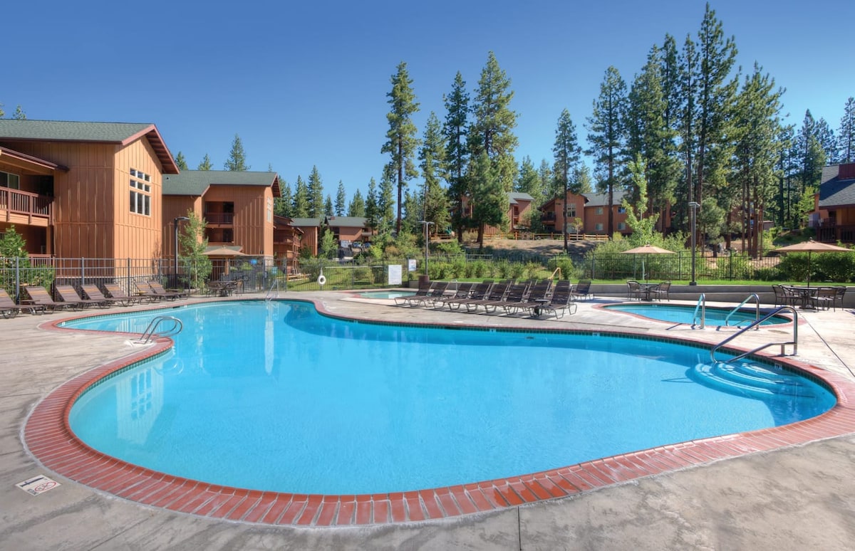 An outdoor swimming pool is surrounded by a spacious deck, featuring multiple lounge chairs arranged nearby. Towering trees and landscaped gardens provide a natural backdrop to the area, enhancing the tranquil environment for relaxation.