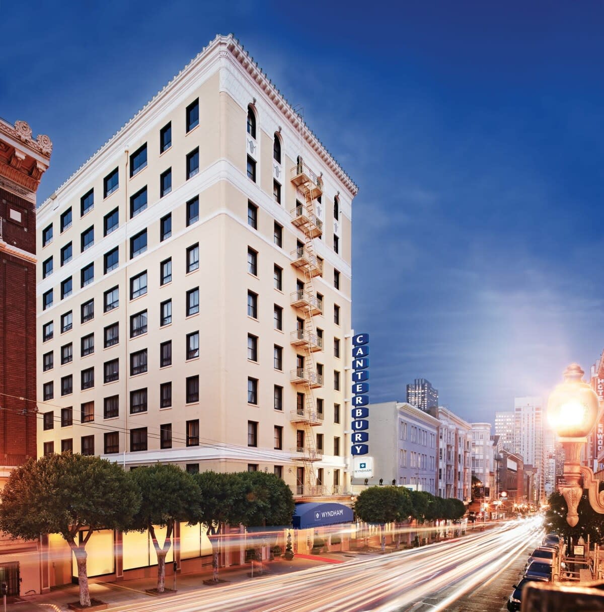 The exterior of the Wyndham Canterbury Resort is shown, featuring a multi-story building with a classic design. Decorative elements and a prominent sign displaying 'CANTERBURY' are visible. Streetlights illuminate the surrounding area, while car lights create streaks along the bustling street.