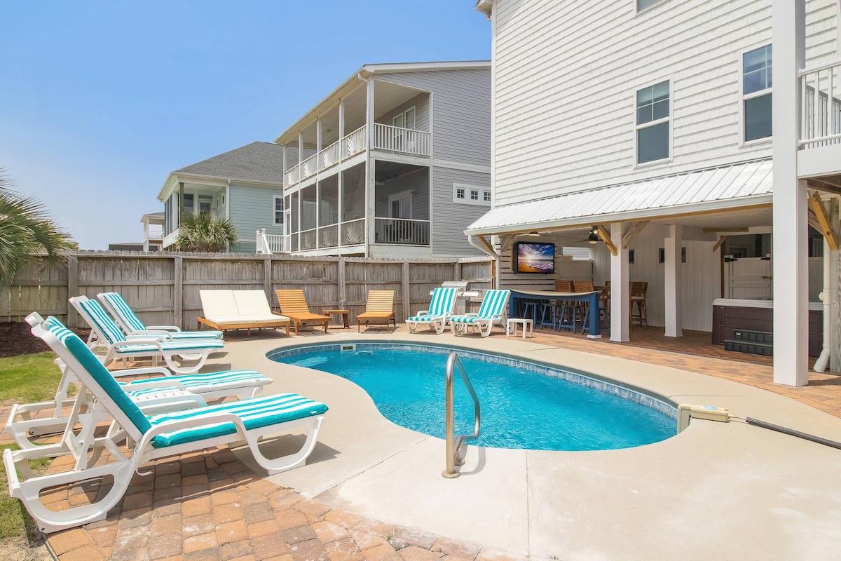 A private pool area features a blue water pool surrounded by lounge chairs and wooden deck furnishings. An outdoor bar area is visible, along with a covered seating space. The enclosed backyard is framed by a wooden fence for added privacy.