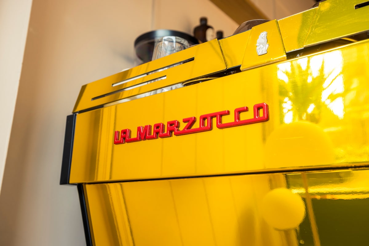 A vibrant yellow espresso machine is prominently displayed, featuring a sleek design with polished surfaces and striking red branding. Glassware is visible on the machine, contributing to a dynamic café atmosphere.