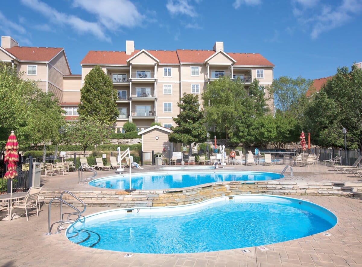 A vibrant outdoor swimming pool area is surrounded by well-maintained landscaping and comfortable lounge chairs. Two interconnected pools, one with a gradual entry, are adjacent to the resort building. Sun umbrellas provide shaded spots for relaxation under the clear blue sky.