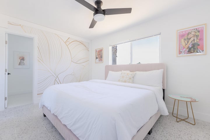 Светла спална соба со чисти бели чаршафи, меки розови детали и природна светлина која создава мирно и просторно место за опуштање.