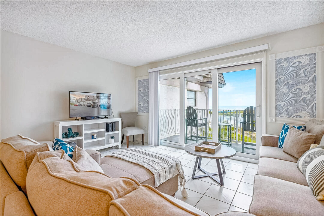 The living area features comfortable sofas arranged around a coffee table, with a large flatscreen Smart TV positioned on a white console. Glass sliding doors lead to the private balcony, showcasing ocean views and allowing natural light to fill the space.