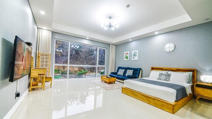 The spacious room features a large bed with a wooden frame and a comfortable blue sofa. A television is mounted on the wall opposite the bed. Natural light fills the space through large windows, revealing a view of the surrounding landscape. Light-colored walls and flooring enhance the bright ambiance.