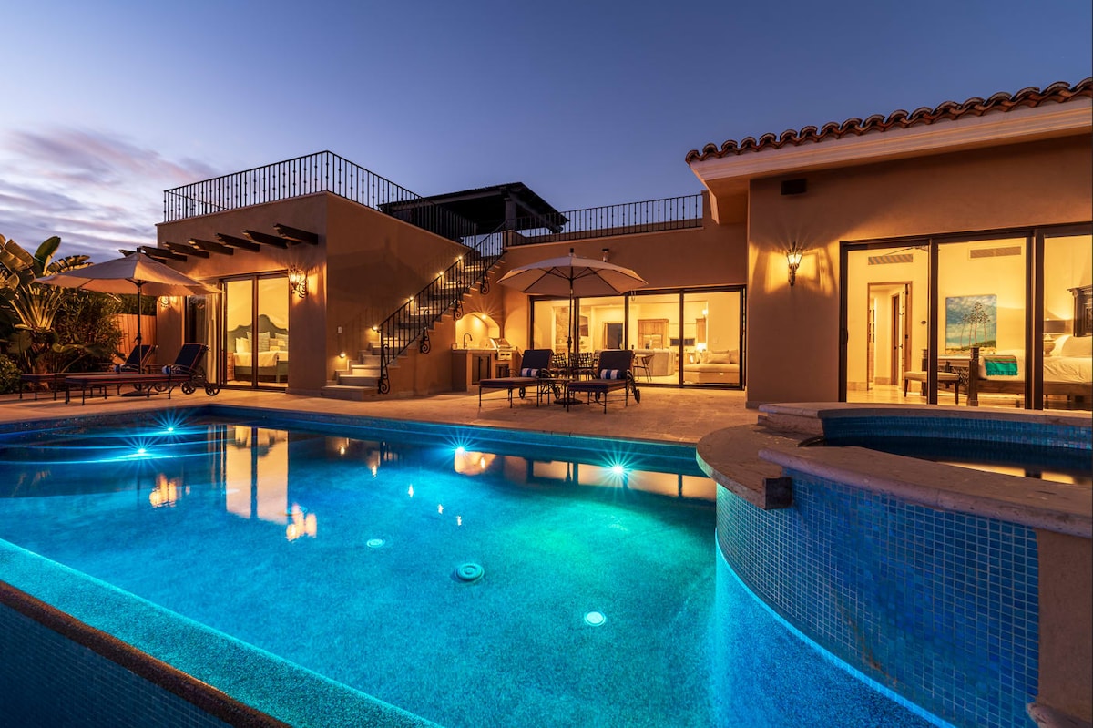 The exterior of the Casita is showcased at twilight, featuring a heated pool illuminated in vibrant blue. The outdoor space includes a cozy seating area and a staircase leading to the rooftop deck. Clear glass doors provide a view into the interior, enhancing the inviting atmosphere.