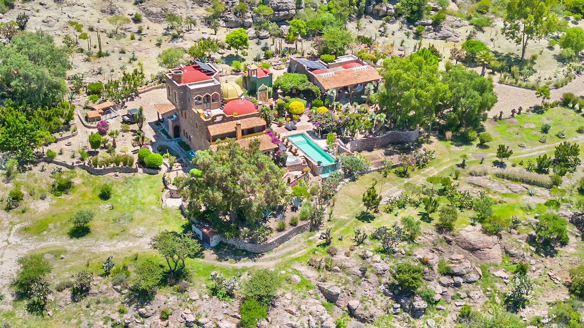 An aerial view captures the stunning layout of Chateau Belle Vue, showcasing lush gardens, an inviting swimming pool, and the unique architectural design of the estate. Surrounding rocky terrain and vibrant greenery frame the property, enhancing its tranquil atmosphere.