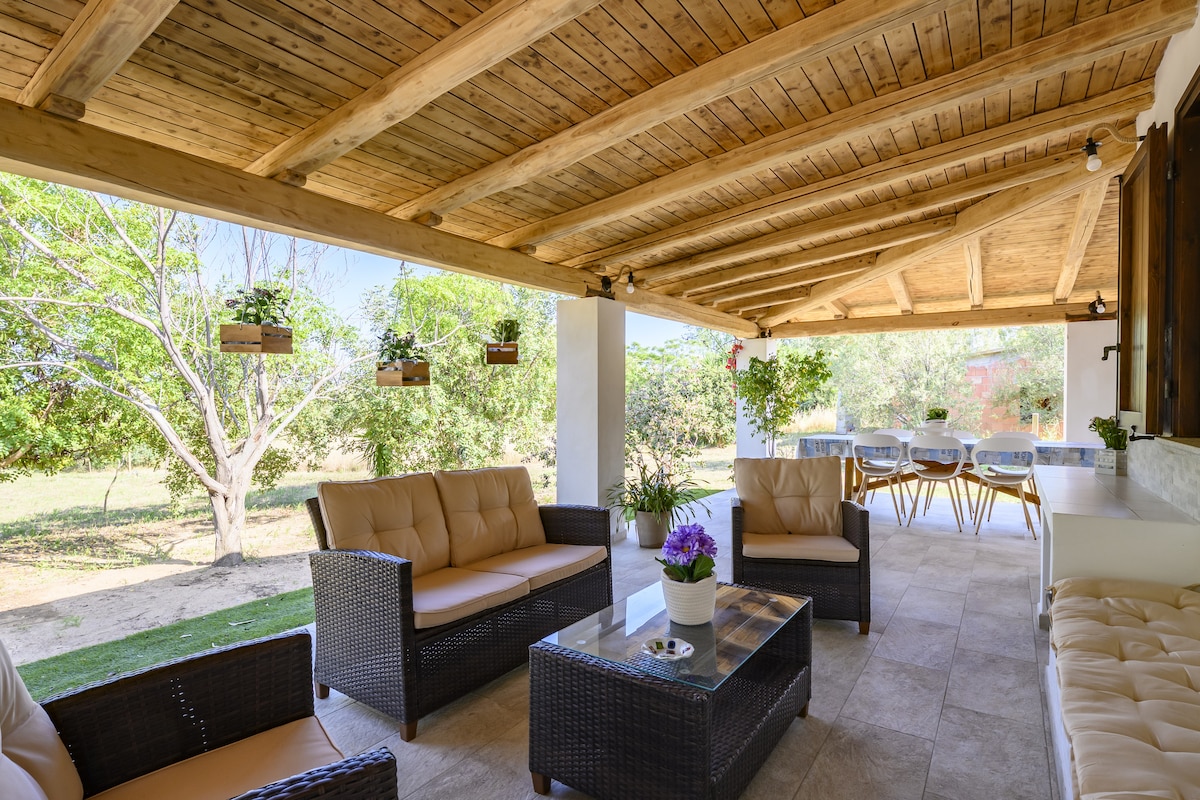 A covered terrace features comfortable seating and a coffee table, creating an inviting space for relaxation. Potted plants hang from the wooden beams, providing natural accents. Beyond the terrace, a lush garden is visible, enhancing the outdoor ambiance.