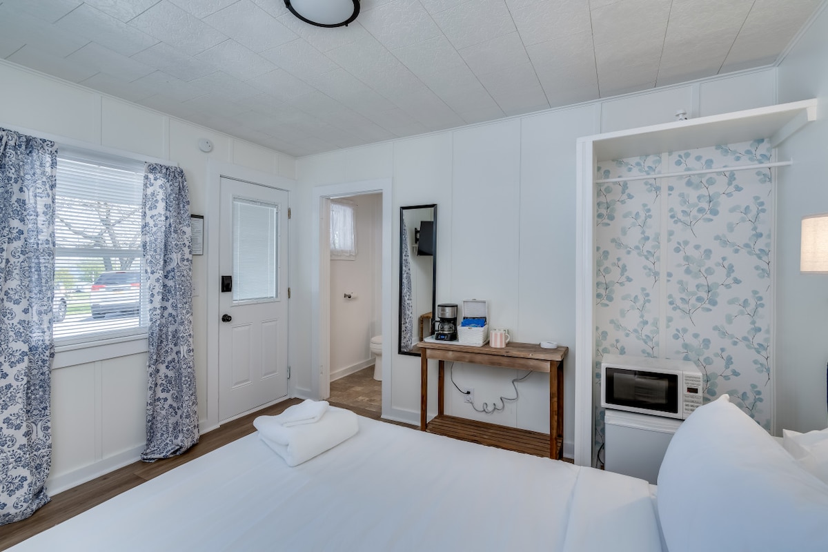 A bright room features a spacious bed with crisp white linens. A wooden table holds a coffee maker and microwave. Floral wallpaper accents a corner, while a large window allows light to fill the space. Dark curtains frame the window, enhancing the calm ambiance.