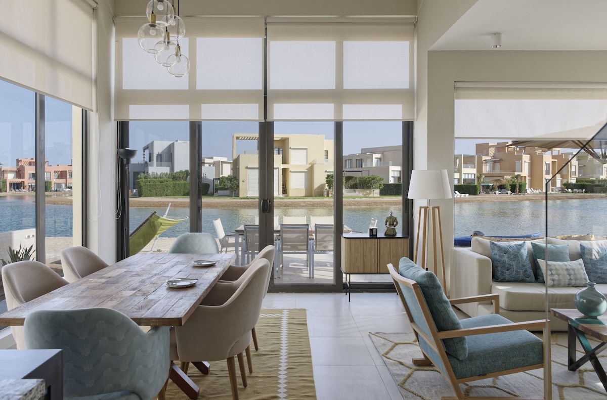 A bright and airy living space features large glass doors that offer views of a serene lagoon. The dining area includes a wooden table surrounded by upholstered chairs, while a comfortable seating area is visible with soft furnishings and a decorative rug.
