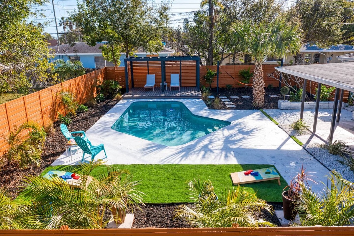 The backyard features an inground pool surrounded by a smooth deck, with lounge chairs positioned beside it. Lush greenery and palm trees are seen throughout the space, enhancing the outdoor living area. A covered pergola provides shade, and a grassy area is included for relaxation.