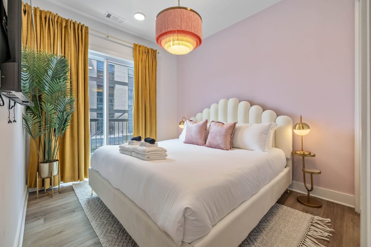 A spacious bedroom is presented with soft pink walls and elegant golden curtains framing a window. The plush bed features a scalloped headboard and is adorned with decorative pillows. A small side table with a lamp complements the cozy ambience, while an indoor plant adds a touch of greenery.