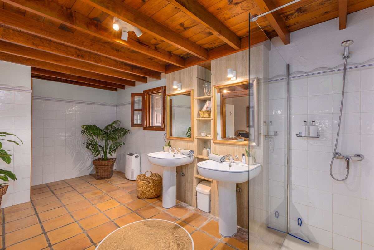 A spacious bathroom features double sinks with mirrored cabinets above. Natural light is provided by a window with wooden frames, and a glass shower is visible. Textured tile flooring and wooden beams overhead create a warm and inviting space, complemented by a decorative plant and woven basket.