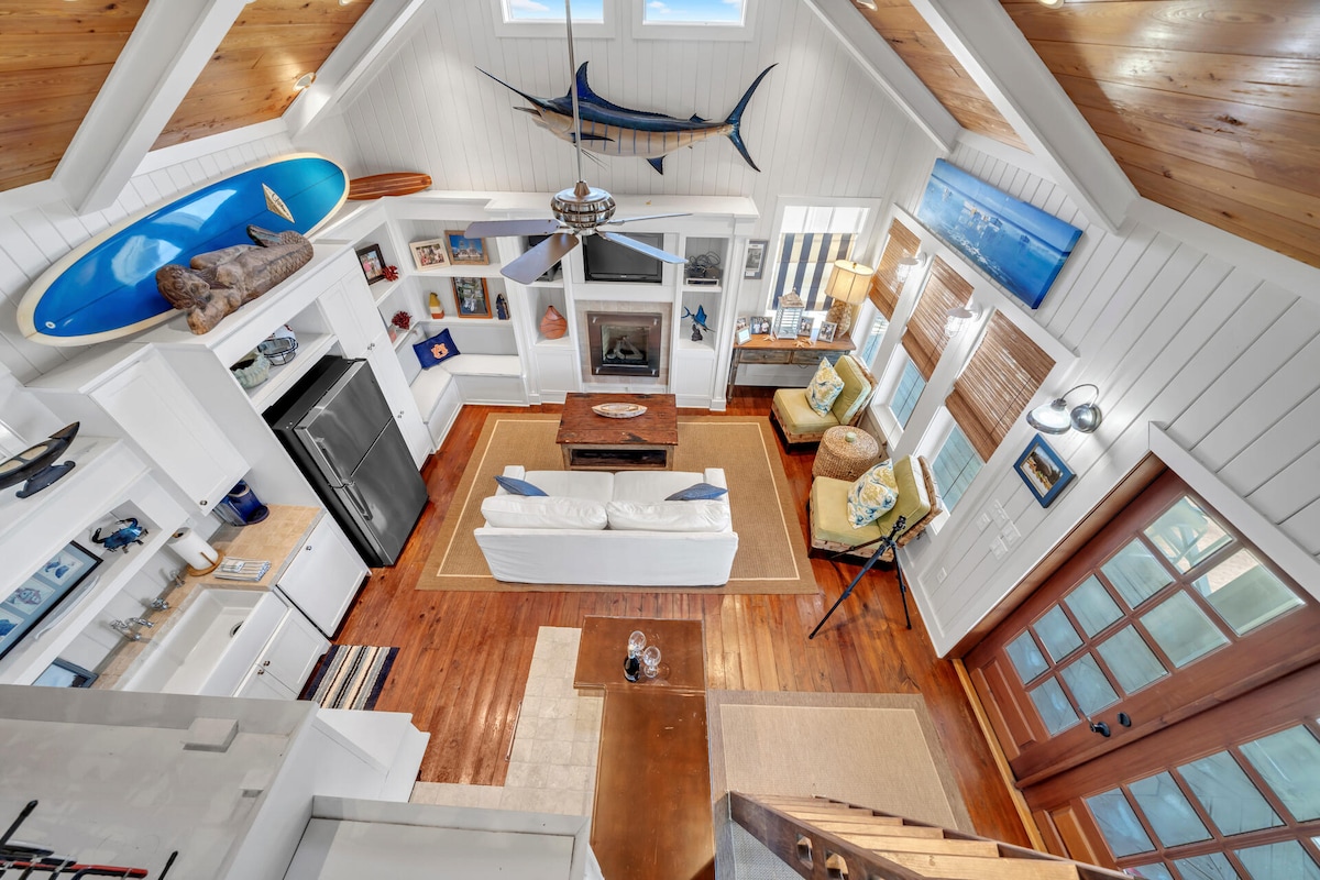 A bright and airy living space is highlighted by high ceilings and large windows. A cozy white sofa faces a central coffee table, while accents of nautical decor are present throughout. A fireplace and blue wall art enhance the comfort of the gathering area.