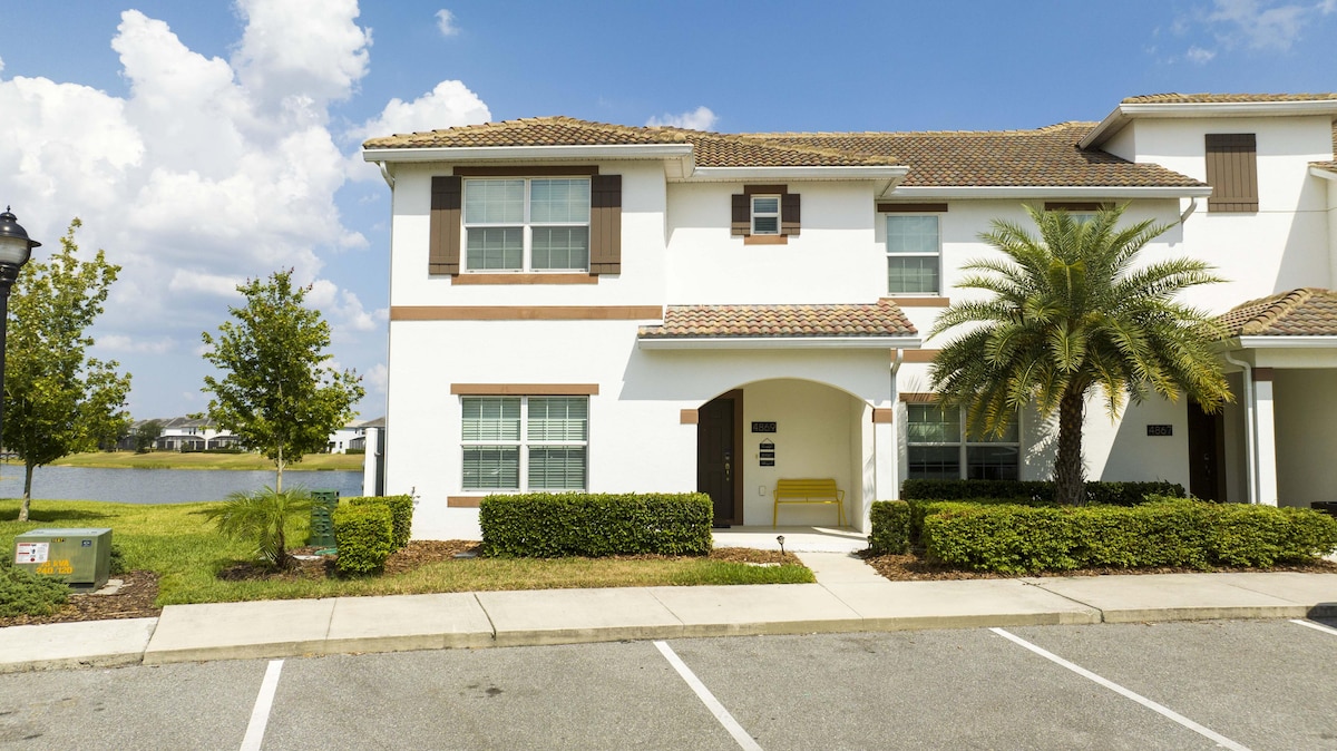 The exterior of a modern townhome is depicted, featuring a well-maintained front yard with shrubs and palm trees. A covered entryway leads into the home, while a tranquil lake is visible in the background, contributing to the serene ambiance.
