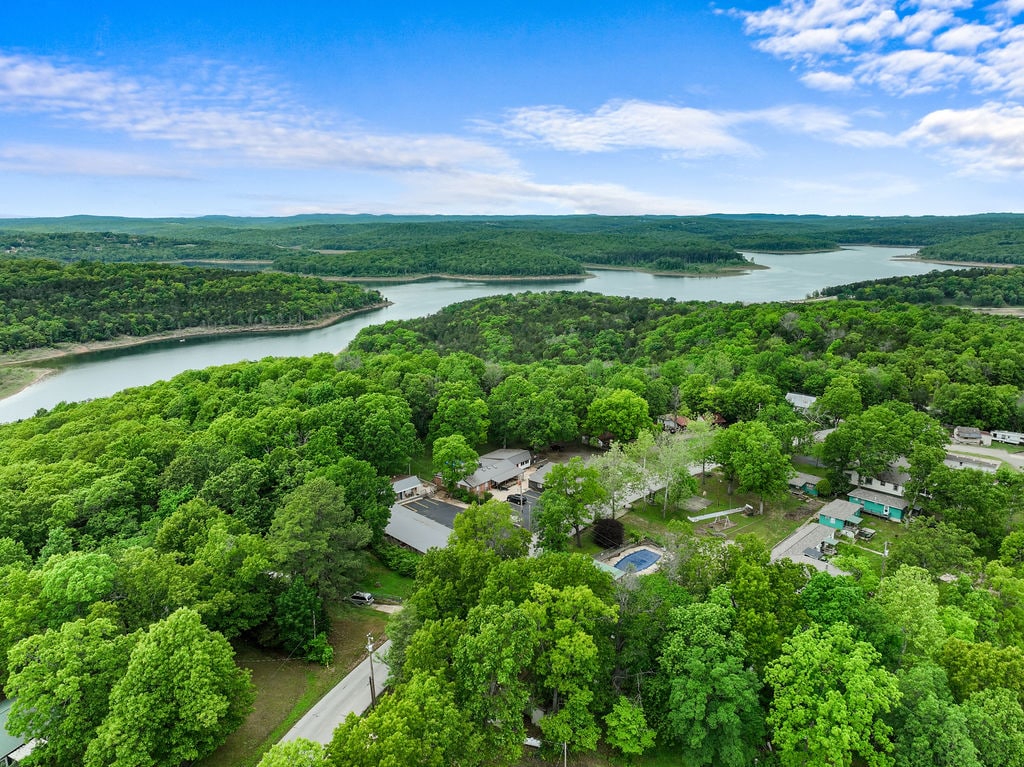 Aerial view reveals the expansive natural landscape surrounding the property, featuring lush green trees and a serene lake winding through the countryside. The scenery highlights the tranquility of the area, with multiple cabins and amenities nestled within the vibrant foliage.
