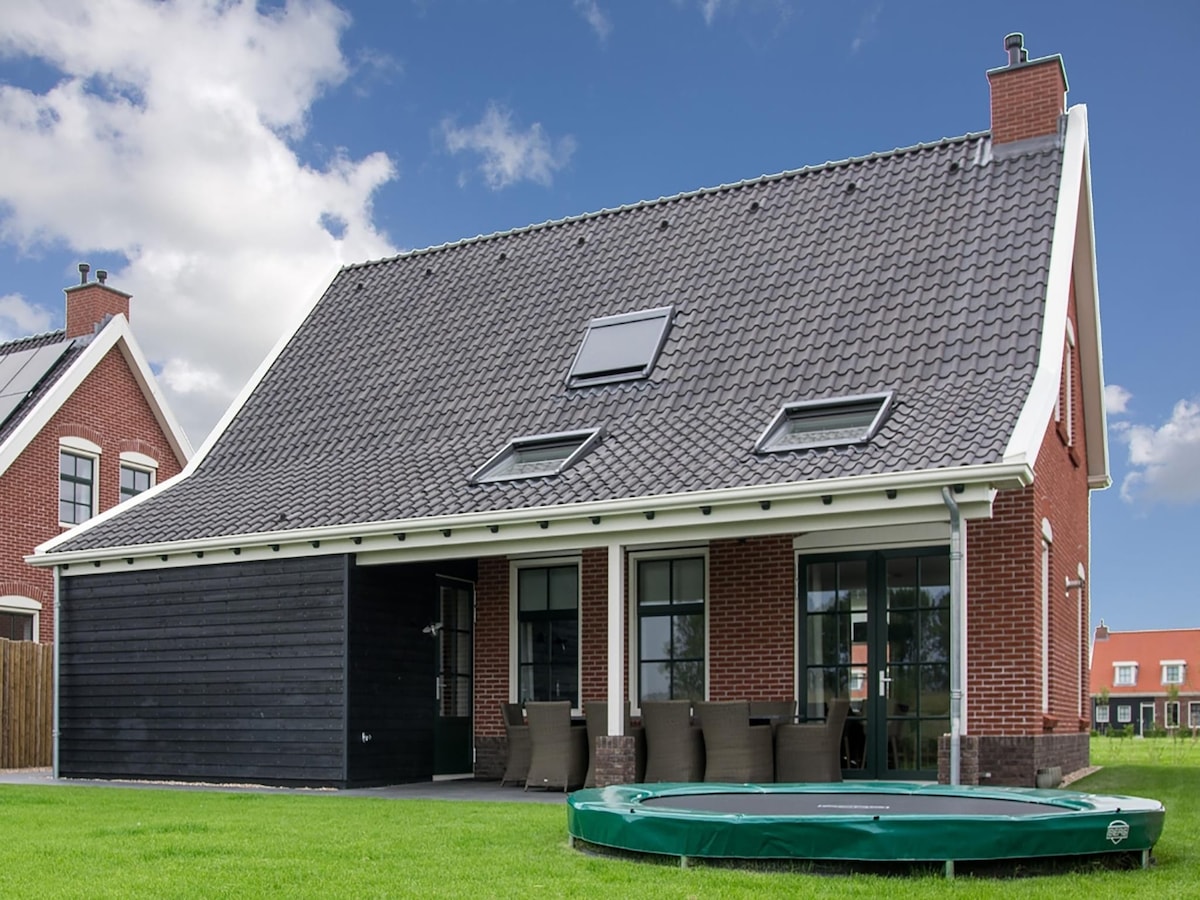 The exterior of the holiday home is showcased, featuring a combination of red brick and dark paneling. A sloped roof with skylights complements the design. A green bubble bath is visible on the lawn, alongside a set of outdoor chairs arranged for relaxation.