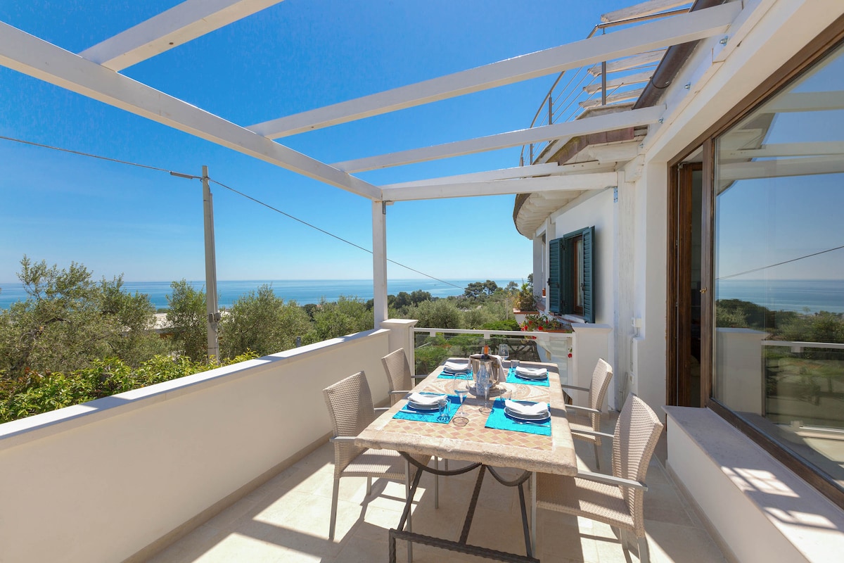 An outdoor dining area is presented under a white pergola, featuring a wooden table set for six. The space overlooks a scenic view of the sea and lush greenery, offering ample natural light and an inviting atmosphere for meals and gatherings.