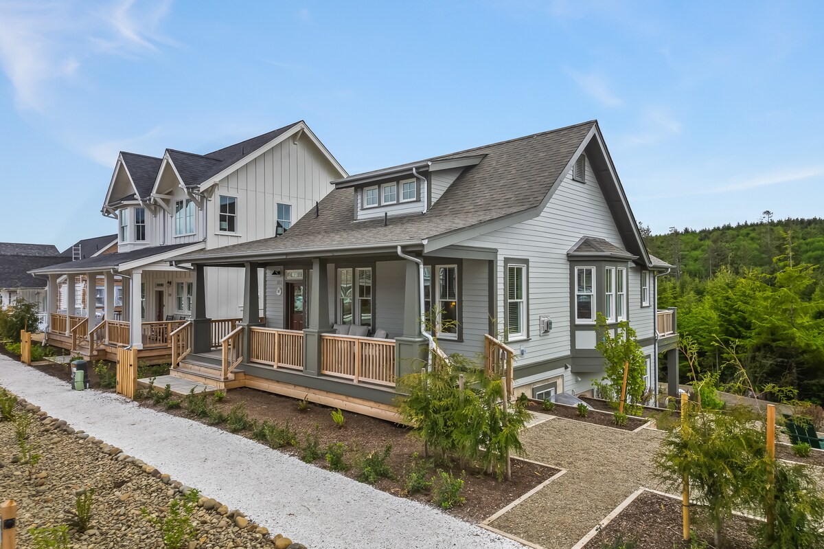 A charming single-story home features a front porch with wooden railings and steps leading to a landscaped pathway. The exterior showcases a mix of clapboard and shingles under a grey roof, complemented by green landscaping and surrounding trees.