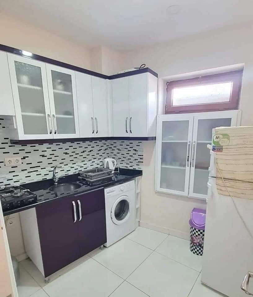 A modern kitchen showcases a combination of white and dark cabinetry. A washing machine is positioned next to a refrigerator. Counter space is available for food preparation, with a gas stove and kettle visible, while a window allows natural light to enter the room.