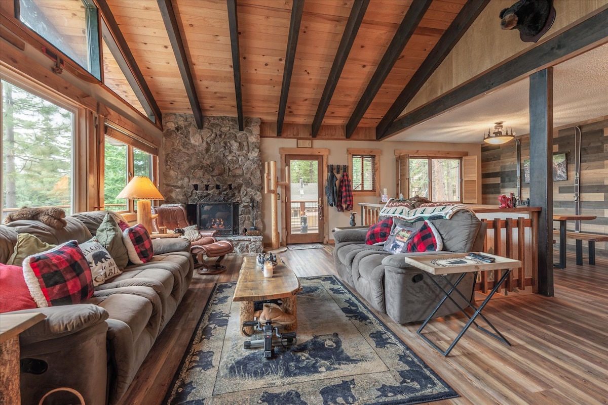 A spacious living area is showcased with a wooden ceiling supported by exposed beams. Comfortable seating is arranged around a wooden coffee table, with a stone fireplace as a focal point. Large windows offer natural light and views of the surrounding trees.