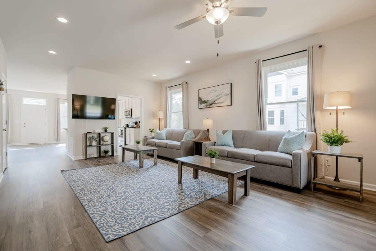 The living room features two comfortable sofa beds across from a SmartTV, with natural light filtering through two windows. A decorative blue area rug is placed on the wood-style flooring, complementing the light walls. A small table with lamps adds functional elegance.