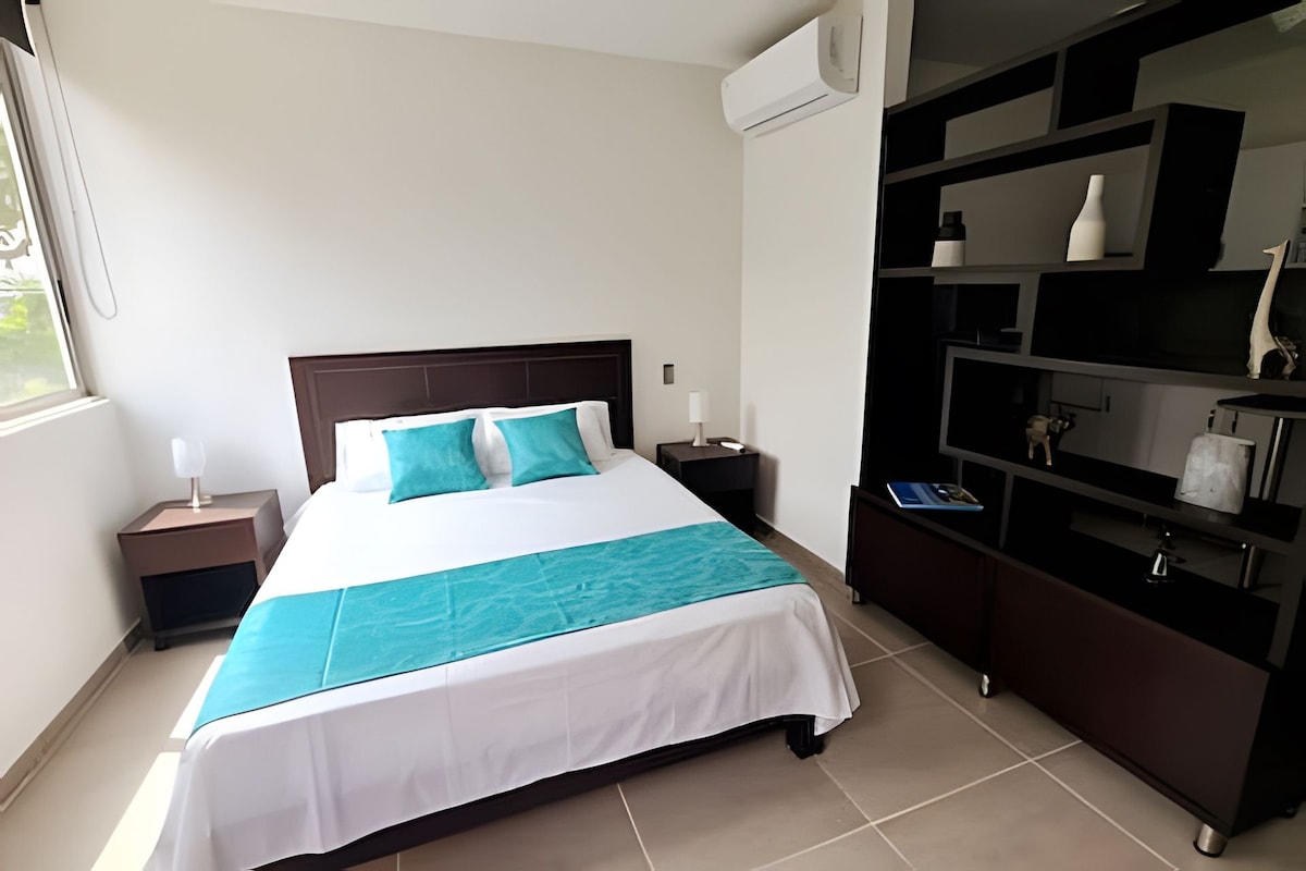 A spacious bedroom features a large bed dressed in white linens with a turquoise accent runner. Nightstands with lamps are positioned on either side. A sleek black shelving unit holds decorative items, while a bright window allows natural light to fill the room.