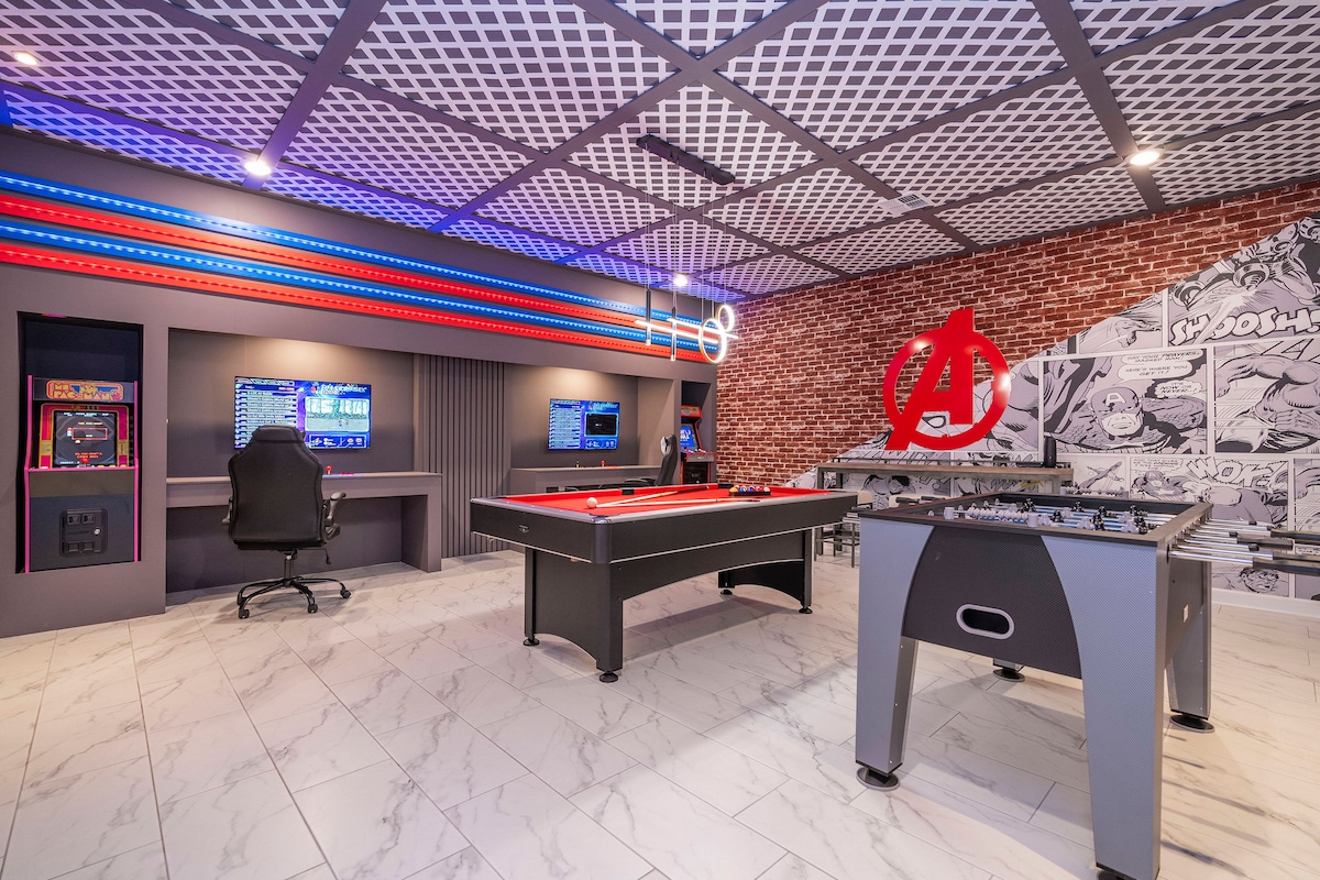 A game room is equipped with a pool table, foosball table, and multiple gaming stations featuring vibrant lighting. A stylish chair sits at a desk, while superhero-themed decor adds a playful touch to the brick wall, creating an engaging atmosphere for leisure activities.