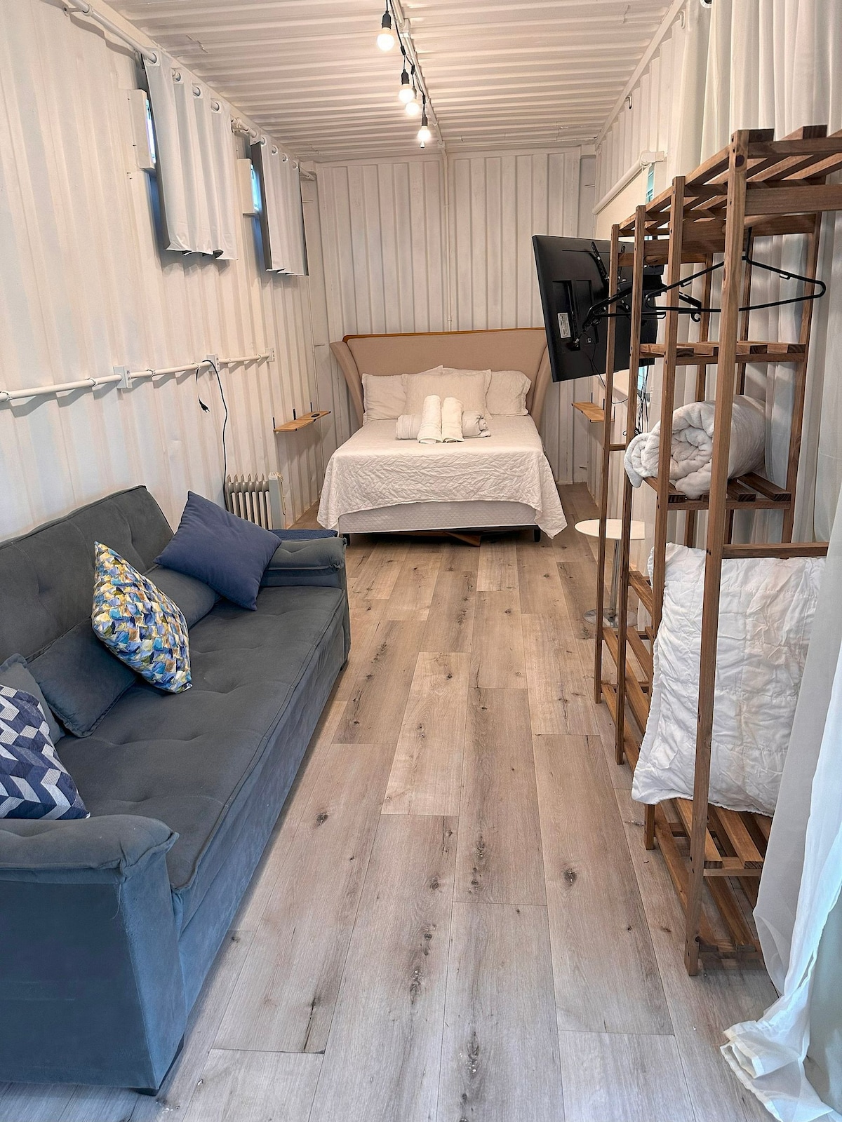 A compact, well-lit space displays a cozy layout featuring a spacious bed with crisp linens. A comfortable sofa accompanies a wooden rack for clothing and extra bedding. Natural light enters through the windows, illuminating the warm wooden flooring and soft wall textures.