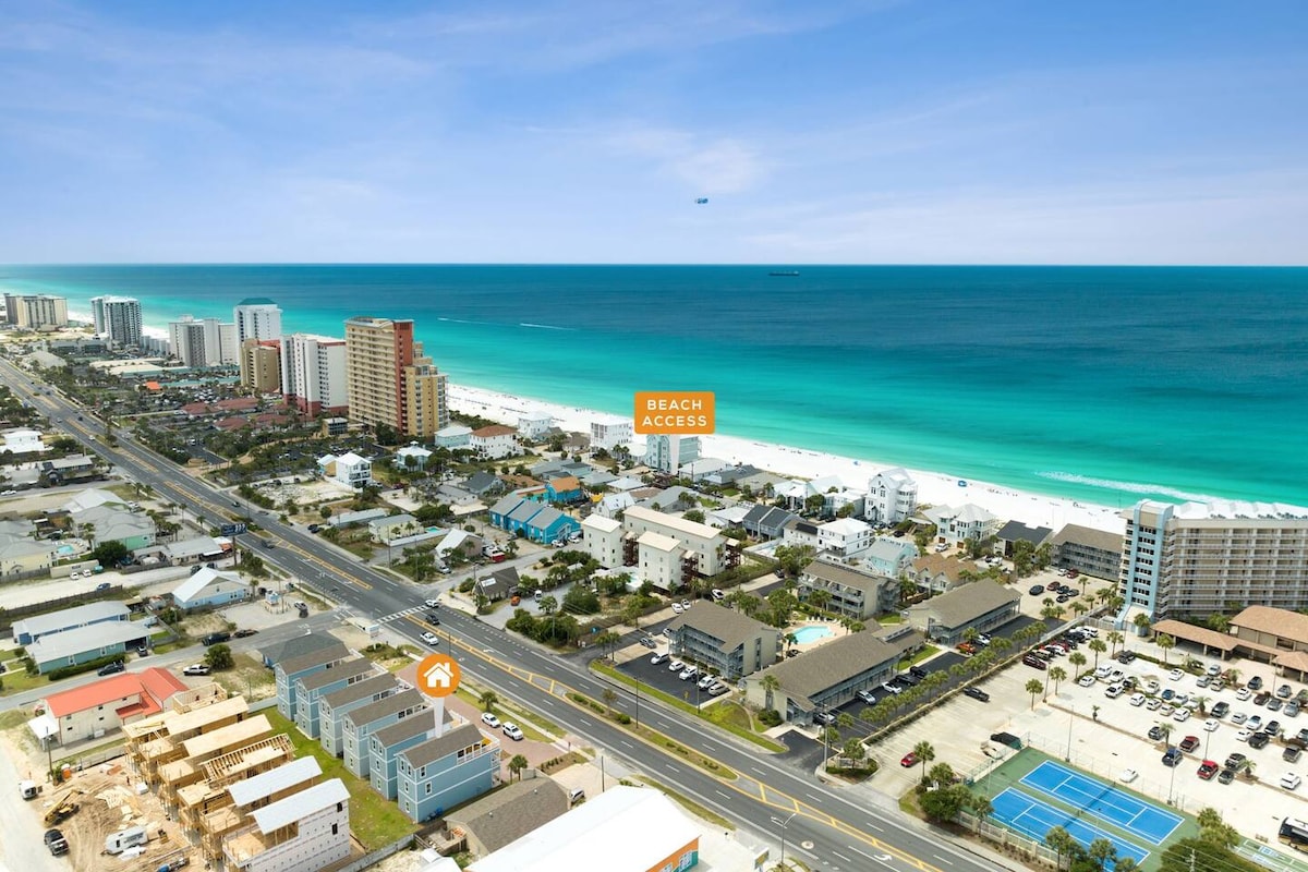 An aerial view captures the vibrant coastline, featuring crystal-clear turquoise waters gently lapping at the white sandy beach. Beach access is prominently marked, with a stretch of beachfront lined by various buildings, including high-rises and vacation homes, creating a scenic coastal atmosphere.