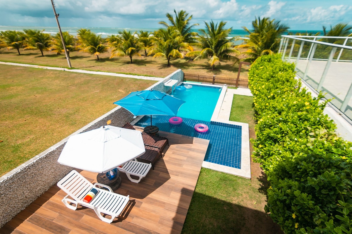 An outdoor area is shown featuring a blue swimming pool surrounded by a well-manicured lawn and palm trees. Sun loungers and umbrellas provide shaded seating, while inflatable flotation devices are positioned in the water, suggesting leisure and relaxation.