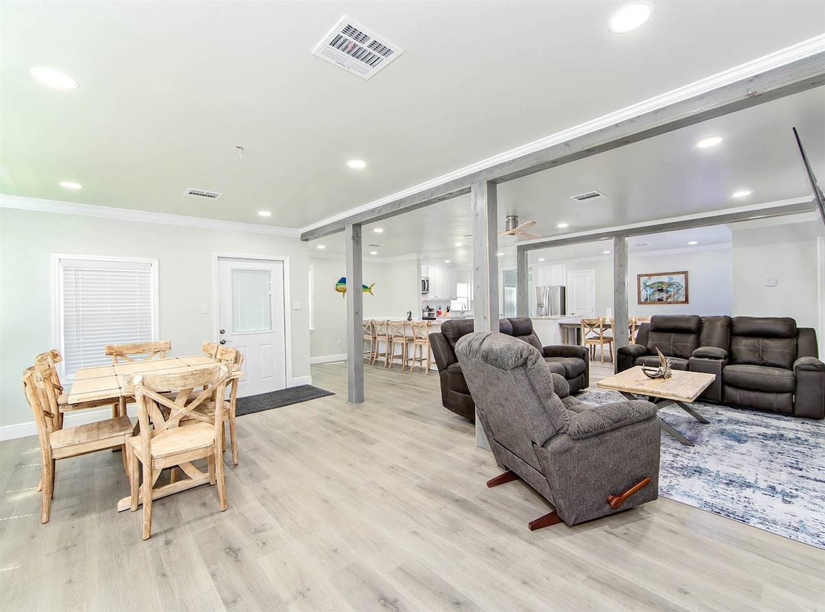 An open living space is captured, featuring a cozy seating area with dark recliners and a light wood dining set. Neutral tones accentuated by soft lighting create an inviting atmosphere. The space seamlessly connects to a kitchen area, enhancing its spacious feel.