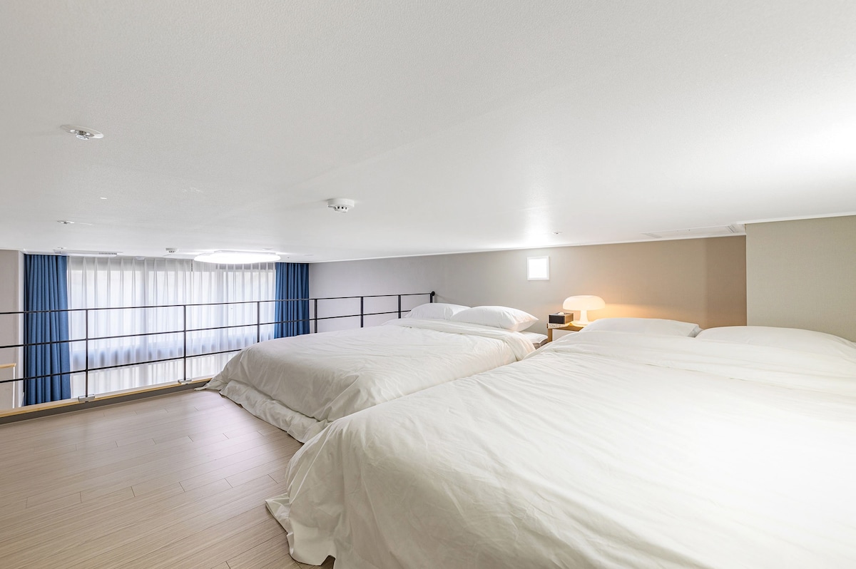 A spacious loft area features two large beds, each dressed in crisp white linens. Natural light enters through a small window, and soft curtains frame the view. A minimalist design with light wood flooring and a simple nightstand enhances the calming atmosphere.