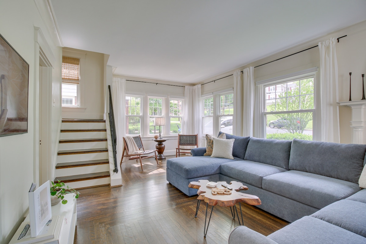 A bright living room features large windows allowing natural light to fill the space. A spacious grey sectional sofa is paired with a wooden coffee table. The room is complemented by light curtains and the staircase leading to the upper level.