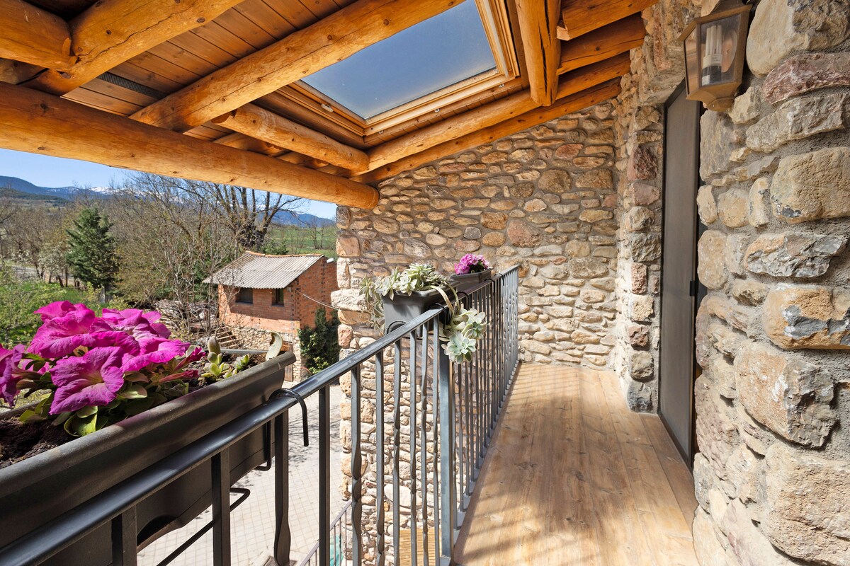 A balcony made of natural wood and stone features a railing and planters filled with vibrant pink flowers. A skylight allows natural light to brighten the area, providing a scenic view of the surrounding landscape and nearby structures.