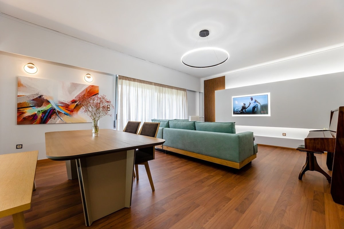 The spacious living area is highlighted by a modern dining table and a comfortable sectional couch. Natural light filters through large windows, illuminating a vibrant artwork on the wall. A sleek flat-screen TV is mounted, and a piano is positioned in the corner, enhancing the inviting ambiance.