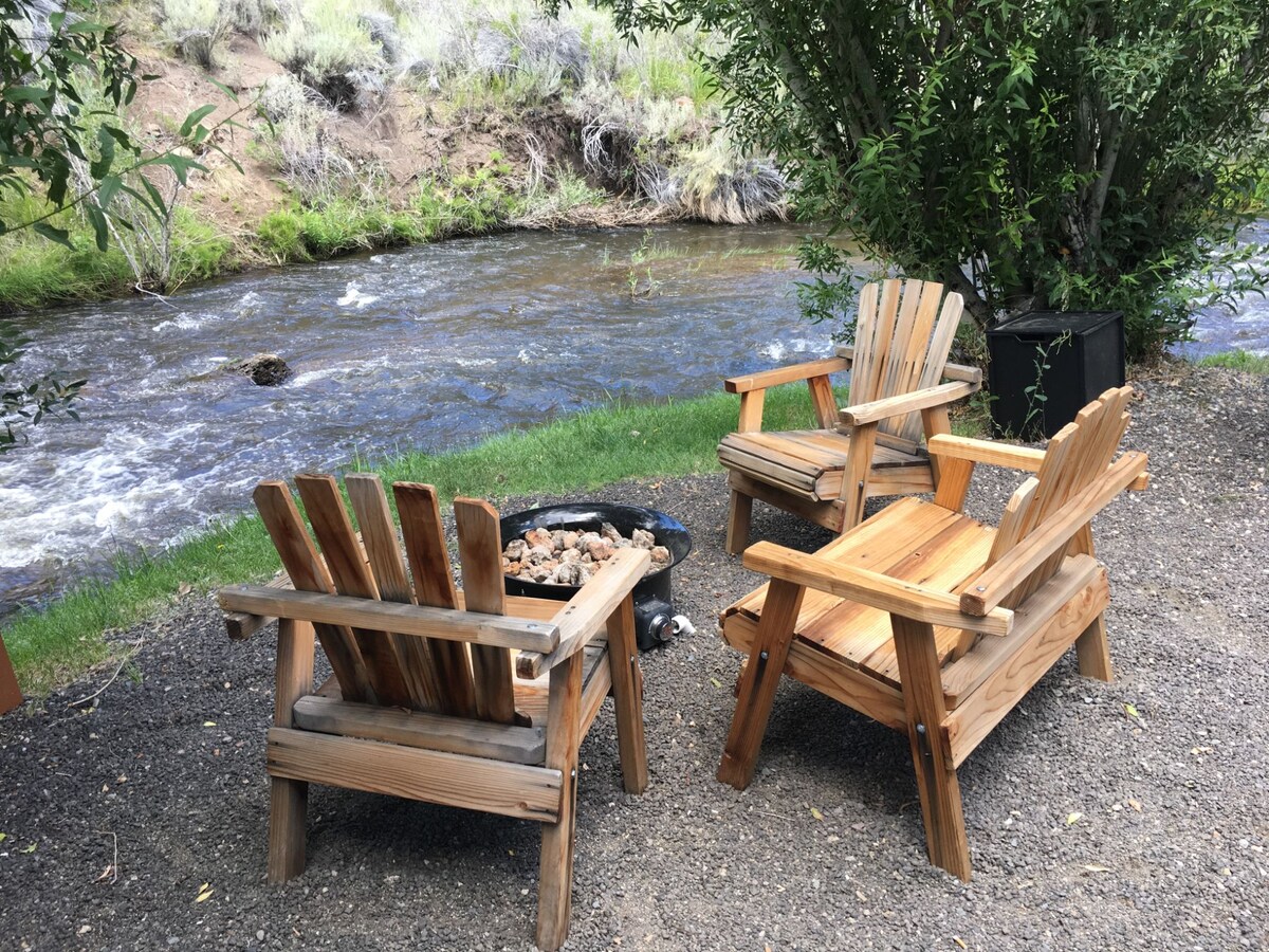 Two wooden chairs with wide slats are positioned beside a flowing creek. A fire ring sits in the center, surrounded by a small pile of rocks. Lush greenery and gentle slopes enhance the serene riverside setting.