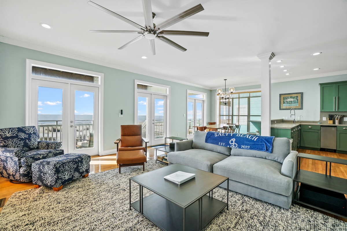 A spacious living area features comfortable seating arrangements, including a sectional sofa and armchair. Large windows provide unobstructed views of the Gulf, while natural light brightens the room. A coffee table is centered on a textured area rug, and a ceiling fan adds to the ambiance.